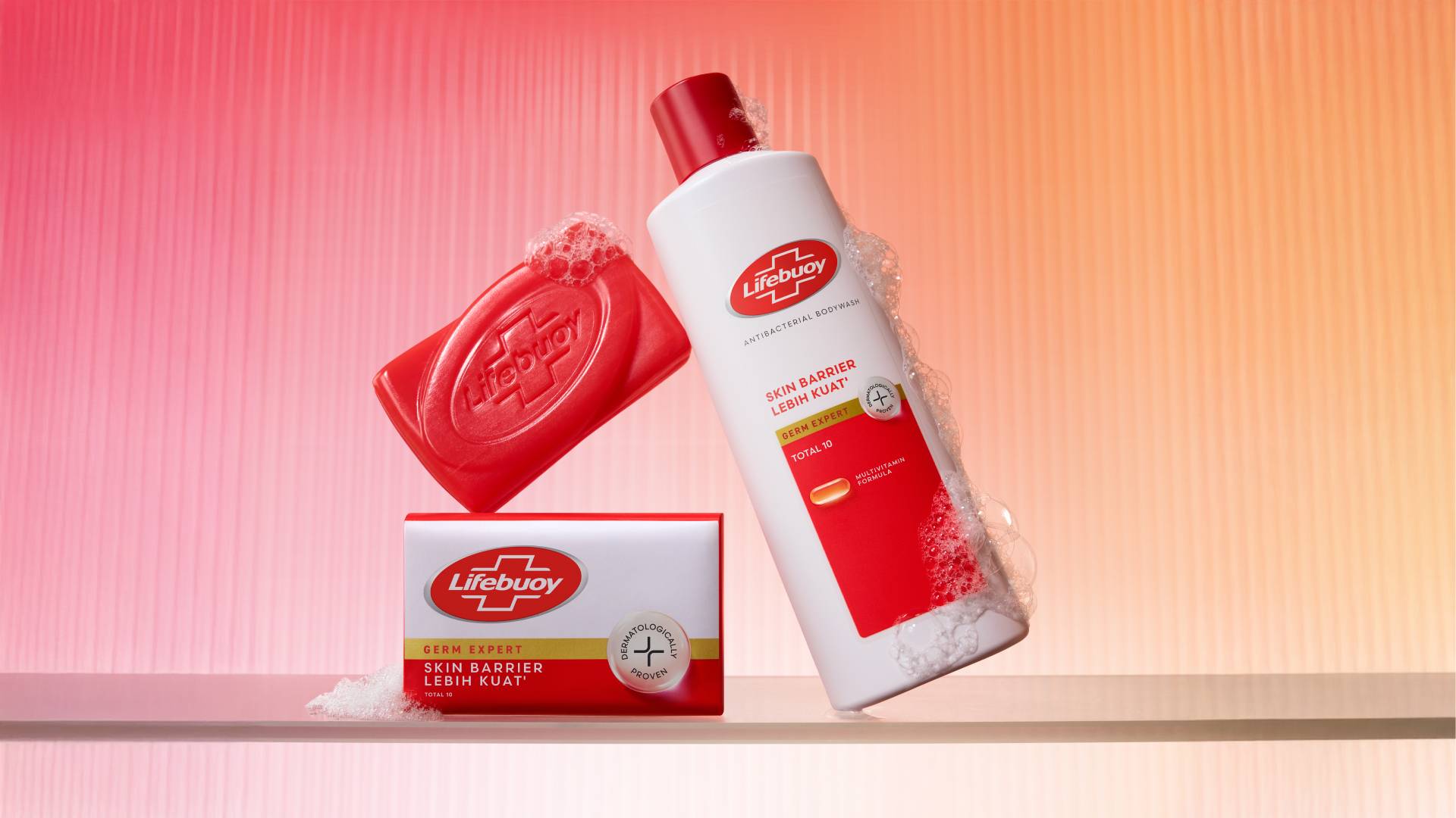Sabun batangan Lifebuoy merah baru dan sabun mandi Lifebuoy baru dalam botol putih, merah, dan emas. Latar belakang berwarna merah muda dan oranye.
