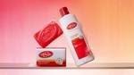 Sabun batangan Lifebuoy merah baru dan sabun mandi Lifebuoy baru dalam botol putih, merah, dan emas. Latar belakang berwarna merah muda dan oranye.