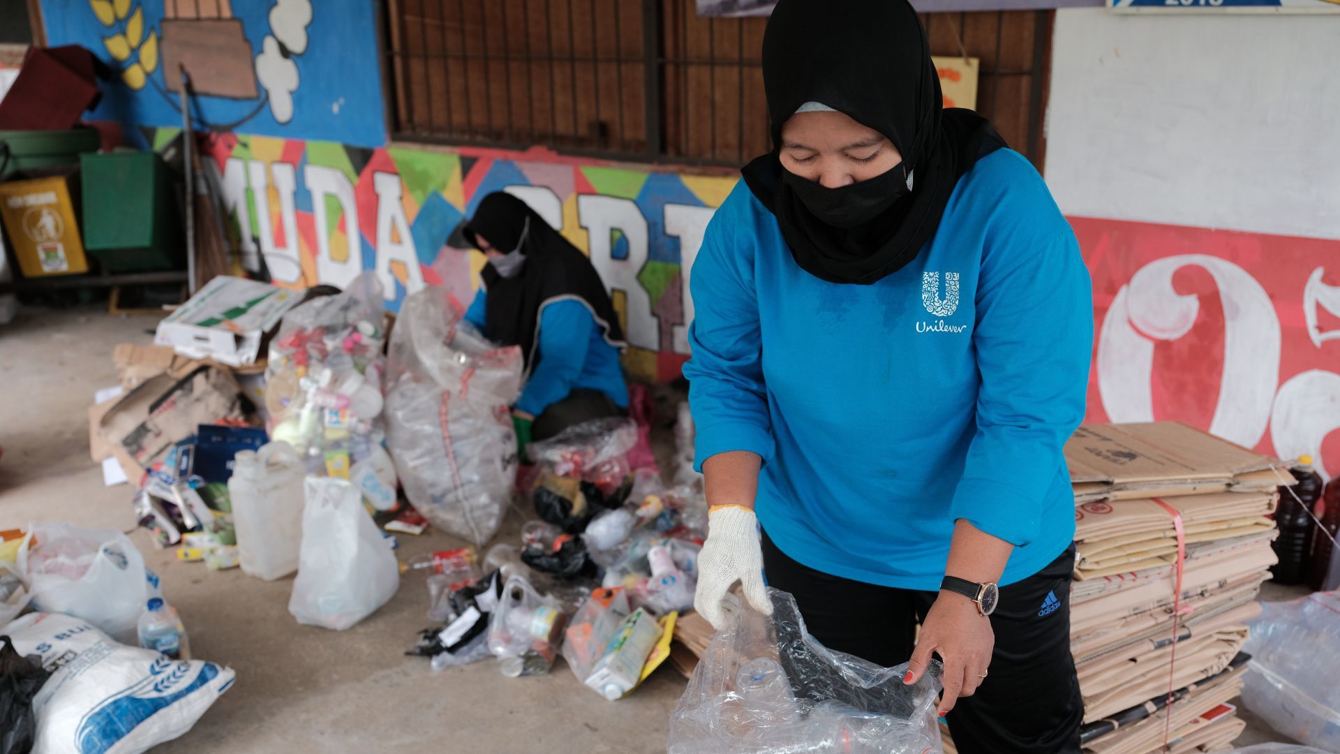 Seseorang berbaju biru memilah sampah plastik di antara tumpukan barang daur ulang di ruang publik yang dicat cerah.