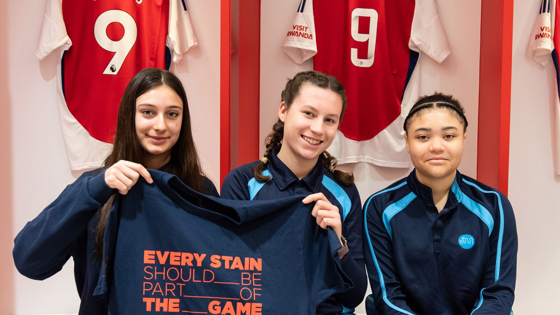 Tiga perempuan duduk di ruang ganti Arsenal FC, memegang kaus bertulis “Setiap noda harus menjadi bagian dari pertandingan”.